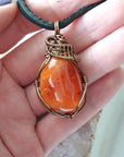 Carnelian pendant for men in hand for size reference – natural orange crystal, bronze copper wire, black leather cord, Virgo birthstone.