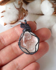 Clear quartz pendant in hand for size reference – natural polished crystal, gunmetal copper wire wrapping, April birthstone.