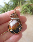Sardonyx pendant in hand for size reference – natural banded stone, bronze and dark silver copper wire, unisex birthstone jewelry by Designs by Nature Gems.