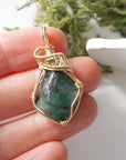 Natural raw emerald pendant shown in hand for size reference, wrapped in detailed 14k gold-filled wire.