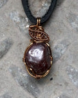 Men’s garnet wire wrapped necklace in antique bronze copper on a black leather cord, shown laying flat to highlight the natural garnet pendant