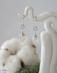 Detailed front view of handcrafted opal sterling silver drop earrings with natural faceted opal, artisan wire wrapping, hanging from a display – Designs by Nature Gems