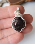 Raw garnet pendant necklace held in hand for size reference, detailed silver plated copper wire wrap