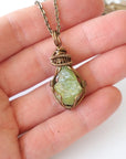Raw peridot pendant for women held in hand for size reference — natural olive green crystal in antique bronze wire wrap, handcrafted August birthstone jewelry