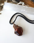 Natural red tiger's eye pendant on black leather cord, laying flat, handcrafted by Designs by Nature Gems.