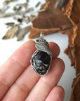 Apache tears obsidian pendant in hand for size reference – natural black volcanic glass, white gold copper wire, unisex jewelry by Designs by Nature Gems.
