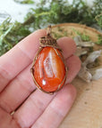 Carnelian pendant in hand for size reference – natural orange crystal, bronze copper wire, unisex Virgo birthstone jewelry by Designs by Nature Gems.