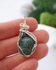 Natural raw emerald pendant shown in hand for size reference, wrapped in detailed soft silver copper wire.
