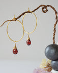 Close-up of handcrafted garnet hoop drop earrings in gold plated brass, featuring wire wrapped natural red garnet gemstones on a jewelry display