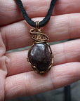 Men’s garnet pendant necklace in antique bronze copper held in hand for size reference, featuring a natural garnet on a black leather cord
