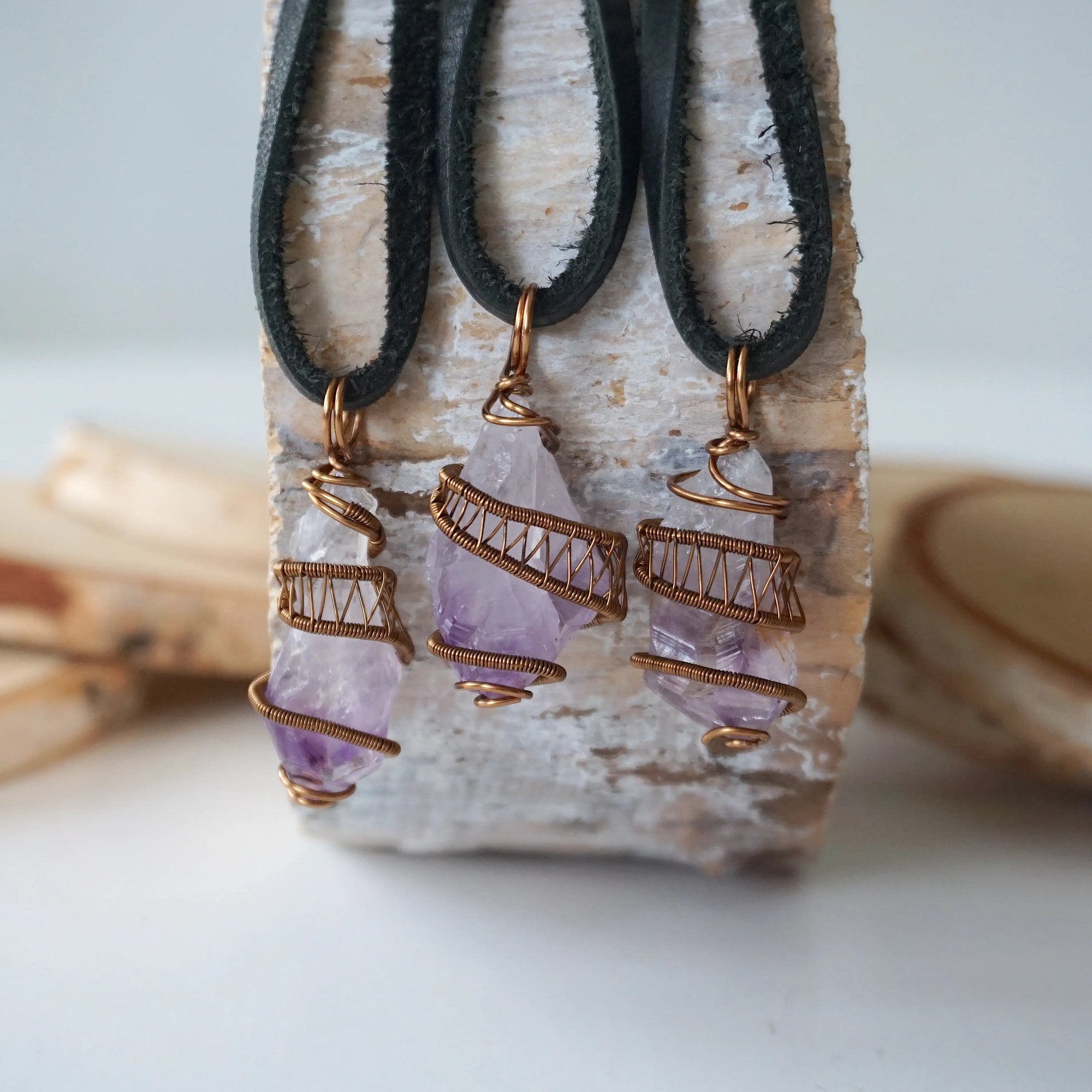 Three raw amethyst pendants on black leather cords showing natural variation in size, shape and purple tone, antique bronze wire wrapped