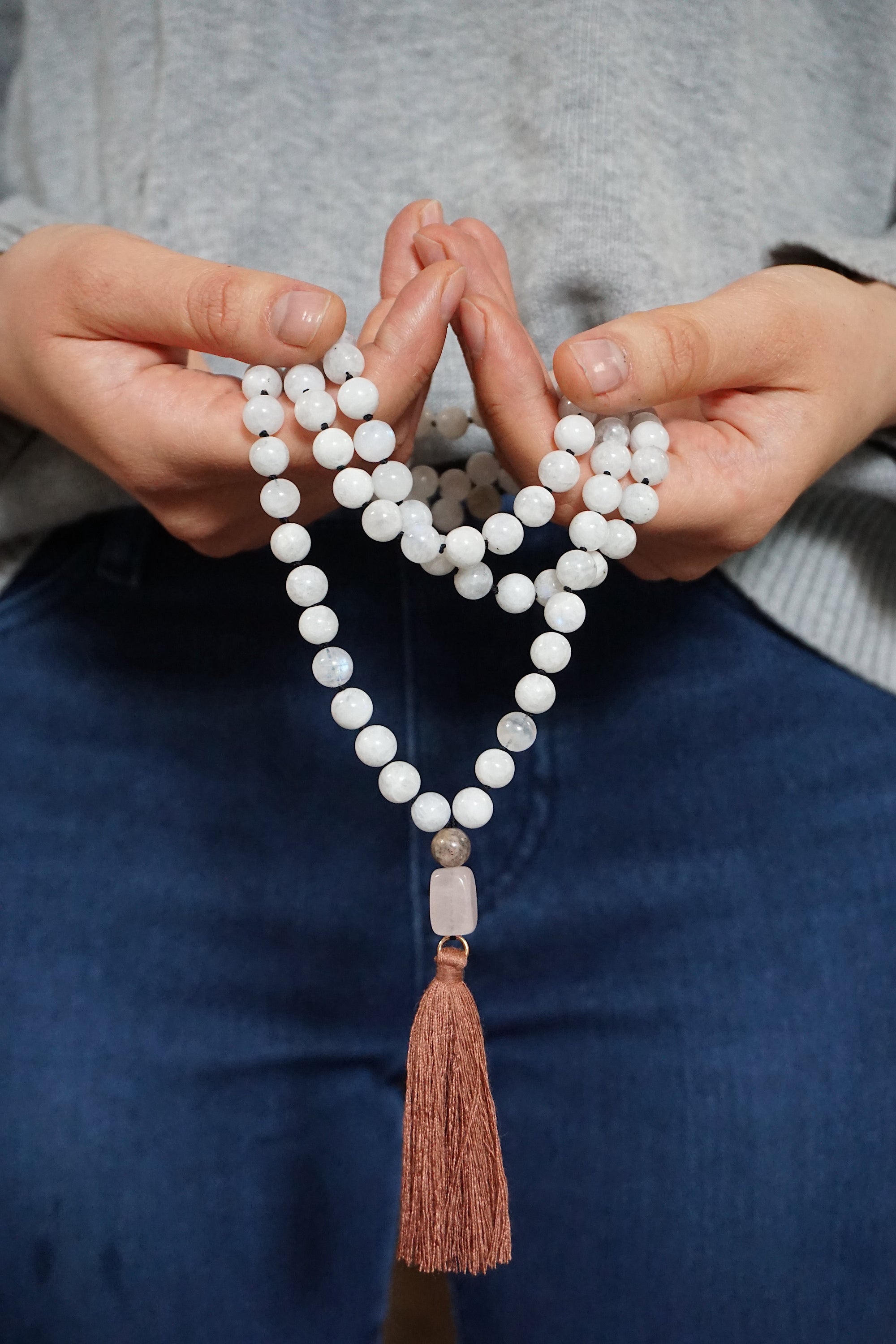 Rainbow moonstone mala necklace with 108 beads and rose quartz guru bead held in hands, knotted silk cord meditation necklace with brown tassel.
