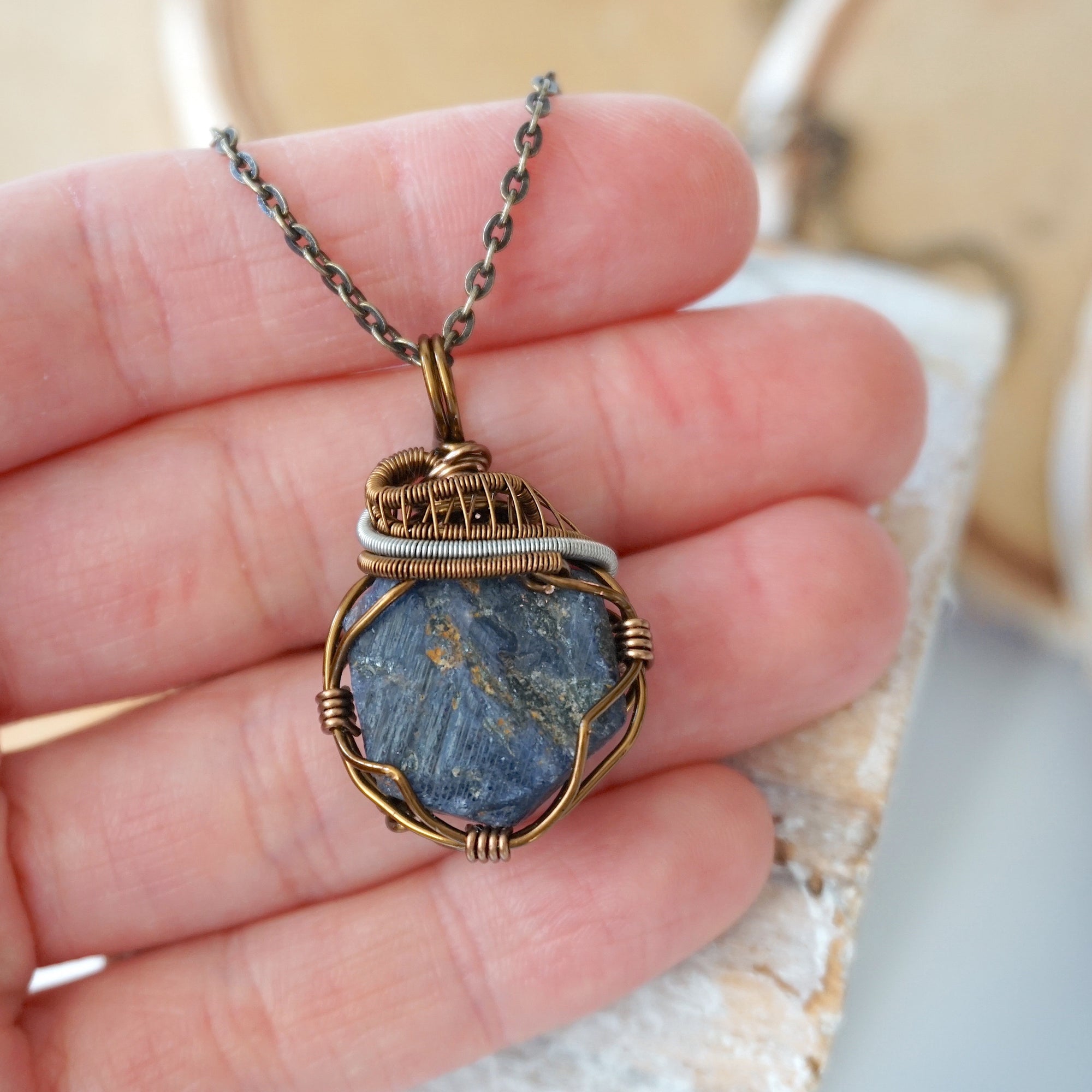 Raw sapphire pendant held in hand to show size — wire wrapped in antique bronze and dark silver, handcrafted crystal necklace by Designs by Nature Gems