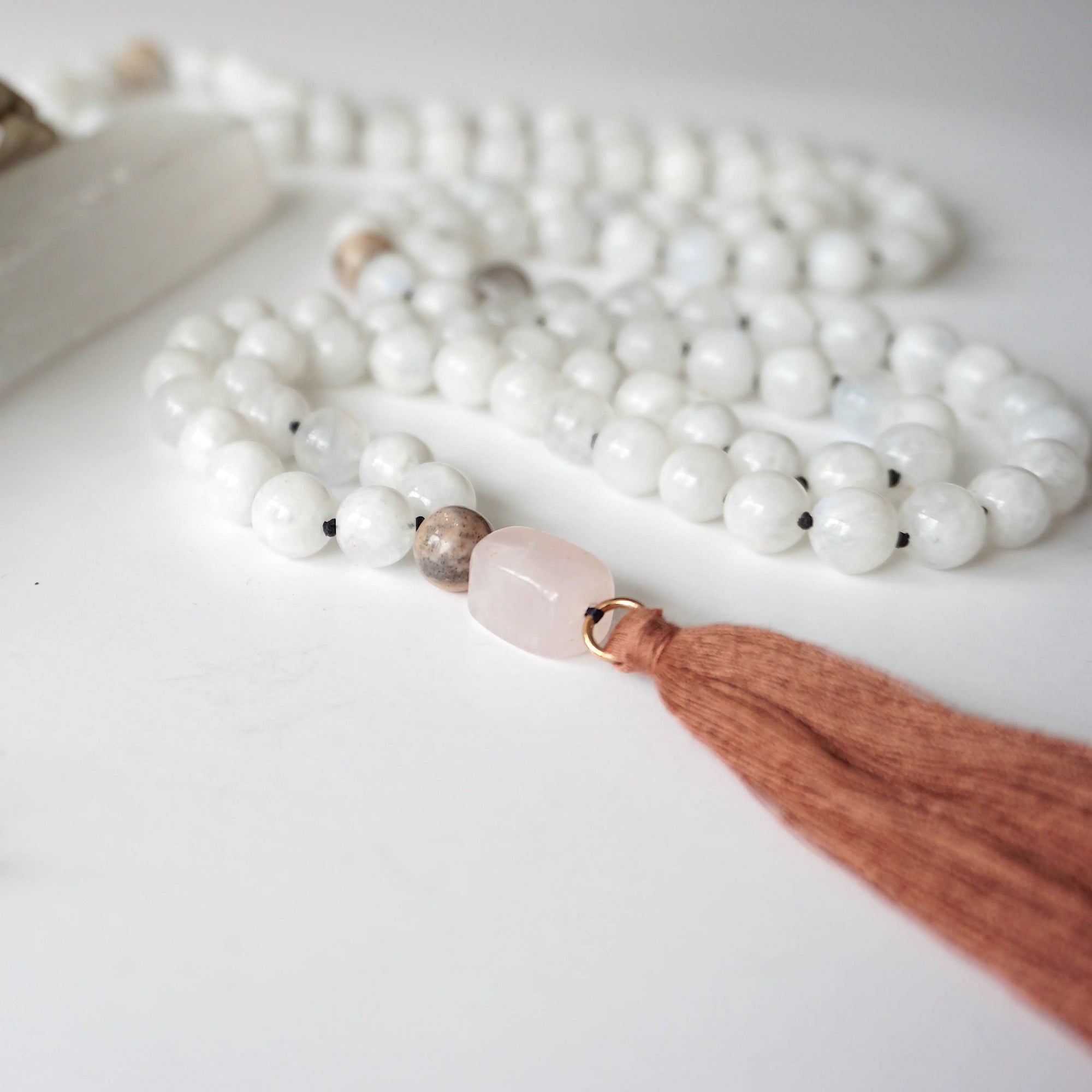 Close-up of rose quartz guru bead and long brown tassel on rainbow moonstone mala necklace, traditional knotted silk cord finish.
