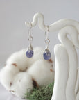 Detailed front view of handcrafted sapphire sterling silver drop earrings with natural faceted sapphire, artisan wire wrapping, hanging from a display – Designs by Nature Gems